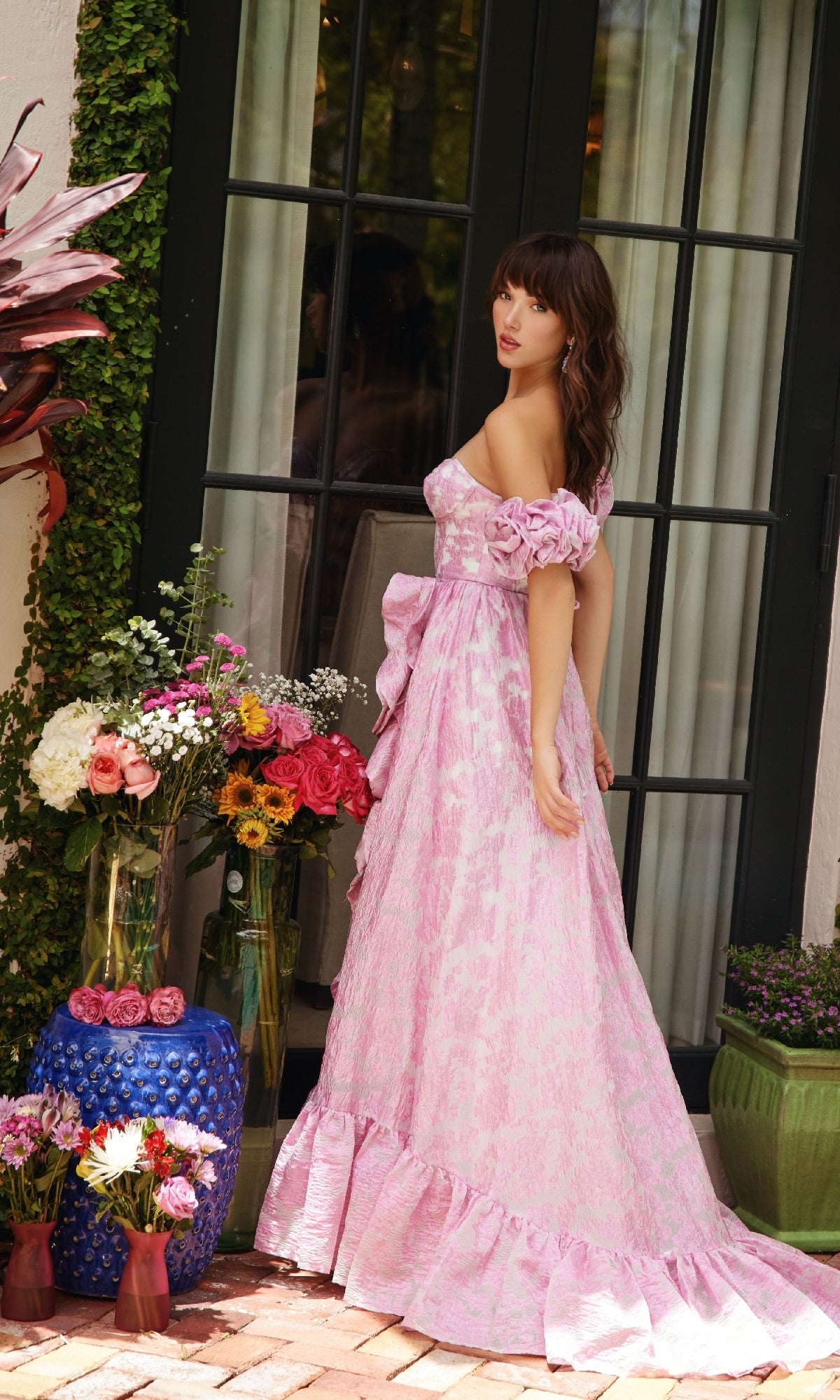 A woman wearing the Ava Presley 40035 long formal dress in pink, featuring an off-the-shoulder ruffled gown with a corset bodice, stands near a glass door surrounded by potted plants and colorful flower arrangements.